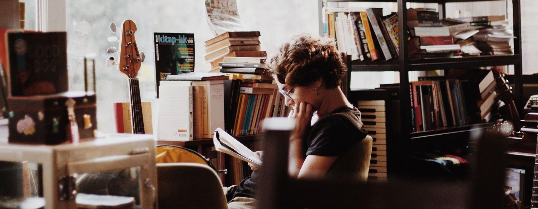 woman, surrounded by books, sits and reads in front of a picture window